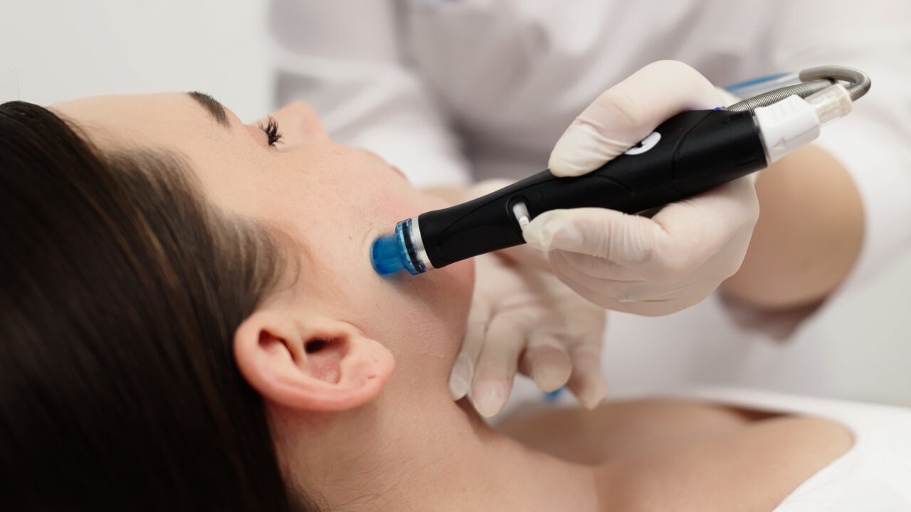 Female patient receiving hydrafacial treatment in a beauty clinic, close-up of facial device held by a technician wearing gloves, soft lighting in the background